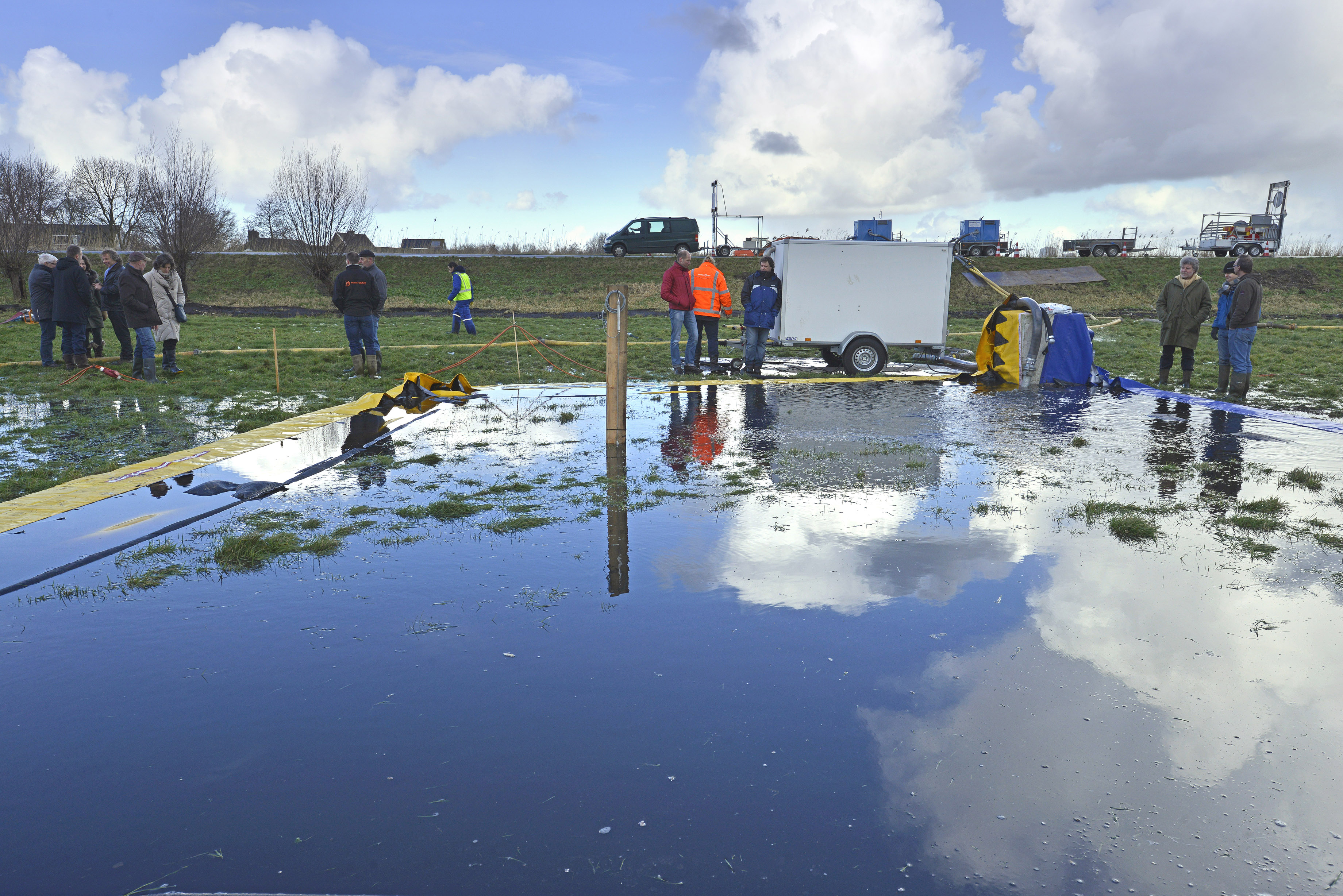 Technici en belangstellenden nemen  tijdens de proef tijdelijke waterkerringen een kijkje bij een ondergelopen stuk weiland