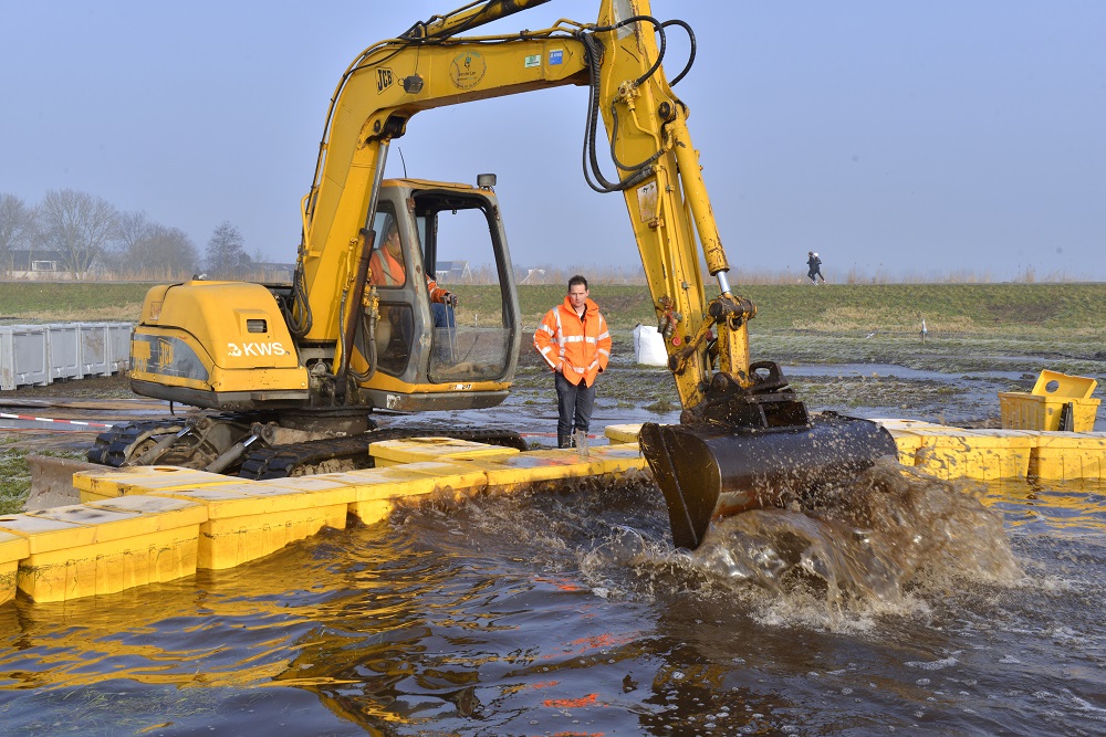 Met een graafmachine wordt een golfproef uitgevoerd in een stuk polder dat voor de pilot is ondergelopen met water